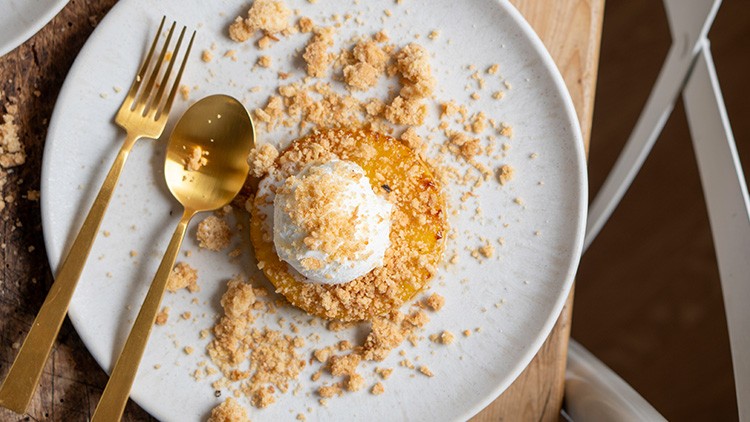 Weiße Porzellanteller mit goldfarbenem Besteck, darauf karamellisierte Frucht, mit weißer Creme und Streuseln garniert.