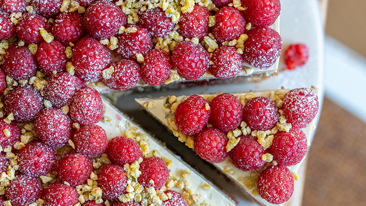 Kuchenstück mit frischen Himbeeren, gehackten Pistazien und Puderzucker auf einer weißen Servierplatte.