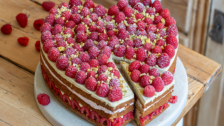 Runder Kuchen mit drei Schichten, frischen Himbeeren, Sahne und zerbröselten gelben Streuseln, ein Stück ist herausgeschnitten.