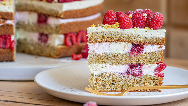 Ein Stück Schichttorte mit Himbeeren, Sahnecreme und Pistazien auf einem weißen Teller mit goldener Gabel.