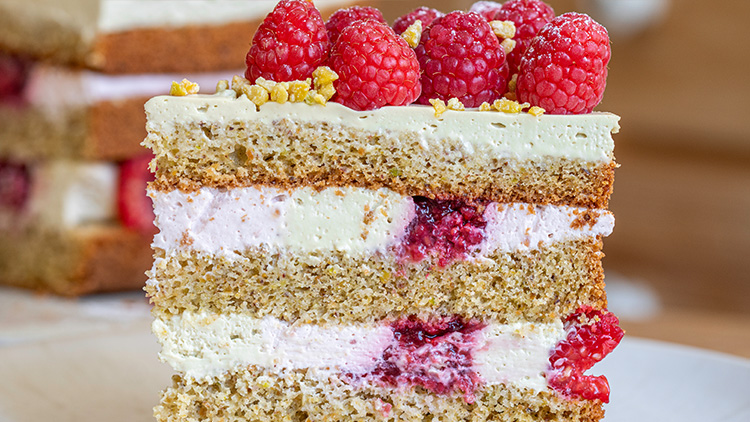 Ein Stück mehrschichtige Torte mit heller Creme, roten Himbeeren und goldenen Streuseln, auf einem Teller serviert.