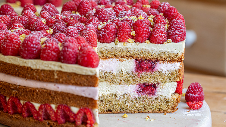 Eine mehrstöckige Torte mit Himbeeren und Sahnefüllung, dekoriert mit frischen Himbeeren und gehackten Nüssen, ein Stück ist herausgeschnitten.