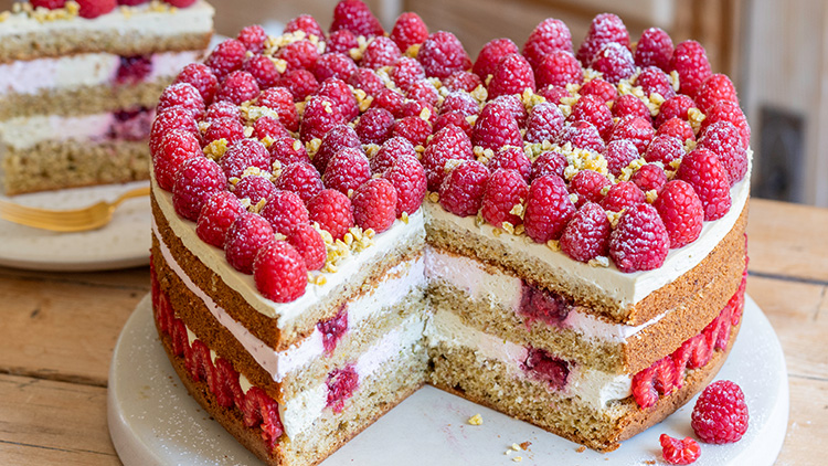 Ein mehrschichtiger Kuchen mit hellem Teig, Sahnefüllung, Himbeeren und gehackten Nüssen darauf, ein Stück ist herausgeschnitten.