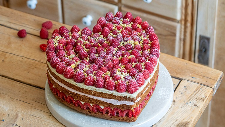 Herztorte aus Biskuit mit Sahnecreme, frischen Himbeeren, gelber Dekoration und Puderzucker auf einem weißen Teller.