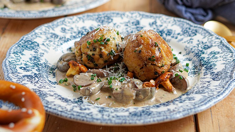 Zwei goldbraune Semmelknödel mit Pilzrahmsauce, gehacktem Schnittlauch und geriebenem Käse auf einem Teller mit blauem Muster.
