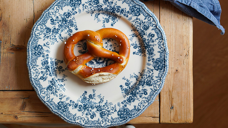 Eine goldbraune Brezel mit Salz liegt auf einem weißen Teller mit blauem Blumenmuster auf einem Holztisch.