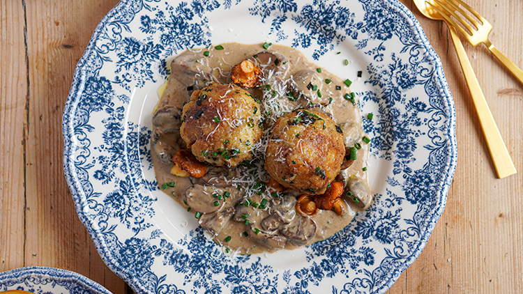Zwei Semmelknödel mit Pilzrahmsoße, geriebenem Käse und Schnittlauch auf einem Teller mit blau-weißem Muster, goldenes Besteck daneben.