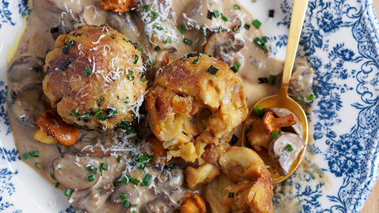 Zwei Semmelknödel liegen in Pilzrahmsoße mit Kräutern und geriebenem Käse auf einem Teller mit blauem Blumenmuster.