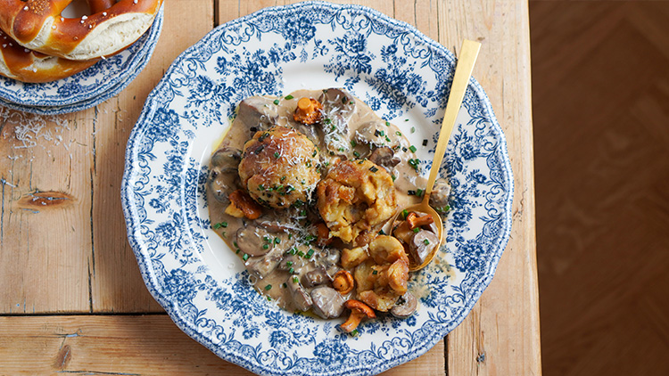 Zwei Knödel mit Pilzrahmsoße und frischen Kräutern auf einem blauen Porzellanteller mit Muster, daneben eine Brezel.