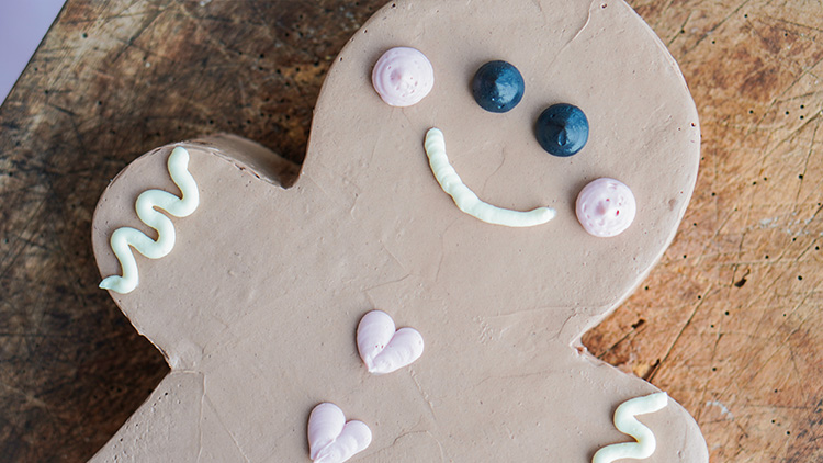 Schokoladenkuchen in Lebkuchenform mit Creme-Dekoration, zwei Blaubeeren als Augen und rosa Herzen auf hellem Holzbrett.