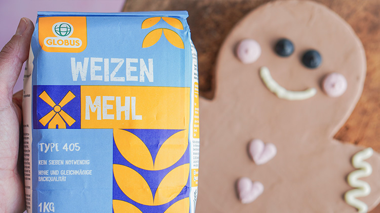 Packung GLOBUS Weizenmehl Type 405 vor brauner Lebkuchenfigur mit Augen, Herzdekor und weißem Lächeln aus Zuckerguss.