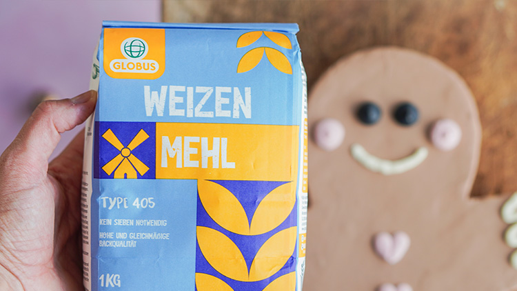 Eine Hand hält eine GLOBUS-Weizenmehl-Packung vor einem Lebkuchenmann mit Zuckerdekoration, Hintergrund ist unscharf.