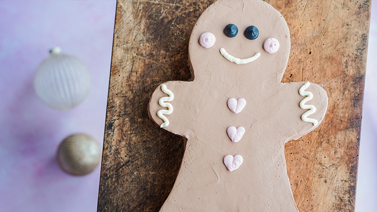Große Lebkuchenfigur mit Augen, Mund und Herzen aus Zuckerdekor auf einem Holzbrett, Hintergrund in Rosa und Beige.
