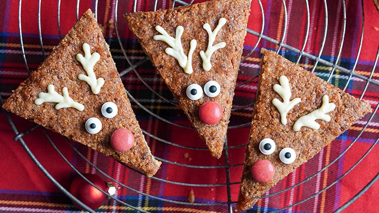 Dreieckige Lebkuchen mit weißen Schoko-Geweihen, Zuckerguss-Augen und roten Nasen auf einem karierten Stoff platziert.