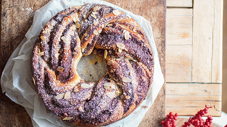 Runder Hefekranz mit dunkler Füllung, bestreut mit Mandelsplittern und Puderzucker, auf Backpapier, Holzuntergrund und roter Deko.