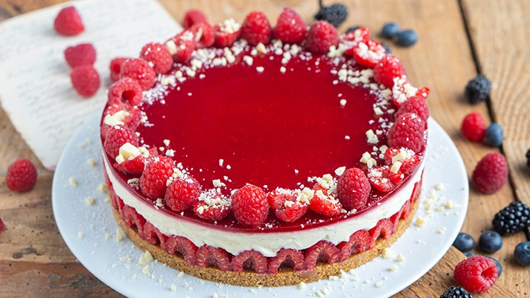 Runde Himbeertorte mit rotem Gelee, weißer Creme und Himbeeren auf einem Holztisch, umgeben von Beeren und einem Notizblatt.