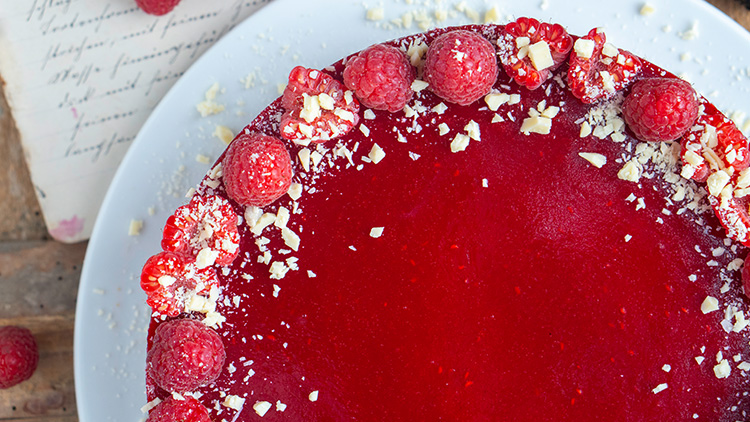 Runde Himbeertorte mit glänzend roter Oberfläche, frischen Himbeeren und weißen Schokoraspeln auf einem weißen Teller.