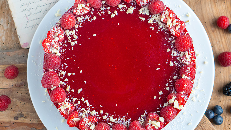 Runder Himbeerkuchen mit roter Glasur, verziert mit frischen Himbeeren und weißen Schokoladenraspeln auf weißem Teller.