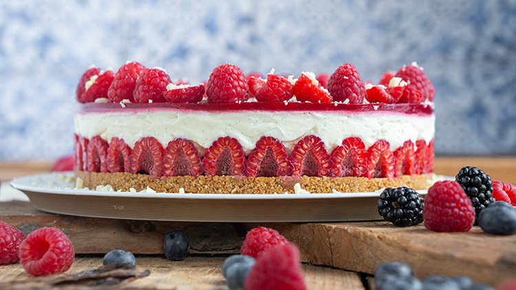 Runde Torte mit Kekskrümelboden, Sahneschicht und Himbeeren, dekoriert mit weißen Schokoladenstücken auf Holztisch.