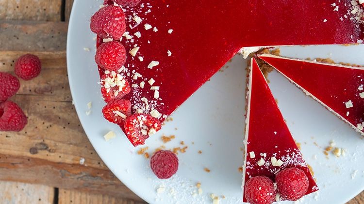 Runde Himbeertorte mit rotem Fruchtspiegel, garniert mit frischen Himbeeren und weißen Schokoladenraspeln, auf weißem Teller.