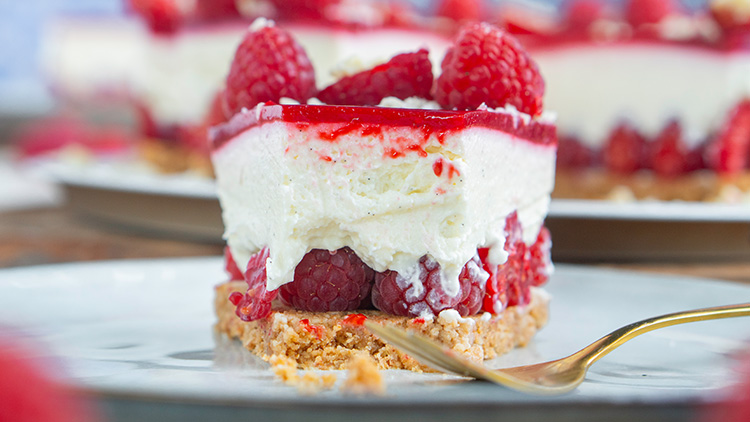 Ein Stück Himbeerdessert mit Keksboden, Sahneschicht, rotem Himbeergelee und frischen Himbeeren auf einem Teller mit goldener Gabel.