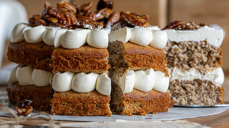 Eine geschichtete Torte aus zwei braunen Böden mit Sahnecreme, dekoriert mit karamellisierten Nüssen, ein Stück herausgeschnitten.