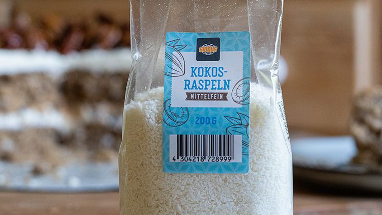 Eine transparente Verpackung mit GLOBUS Kokos-Raspeln, mittelfein, 200 g, und sichtbarem Barcode steht auf einem Holztisch.