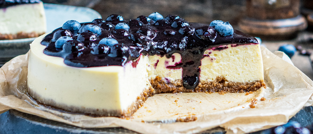 Ein angeschnittener Käsekuchen mit braunem Boden und Blaubeerdeckschicht auf braunem Backpapier, dekoriert mit frischen Blaubeeren.