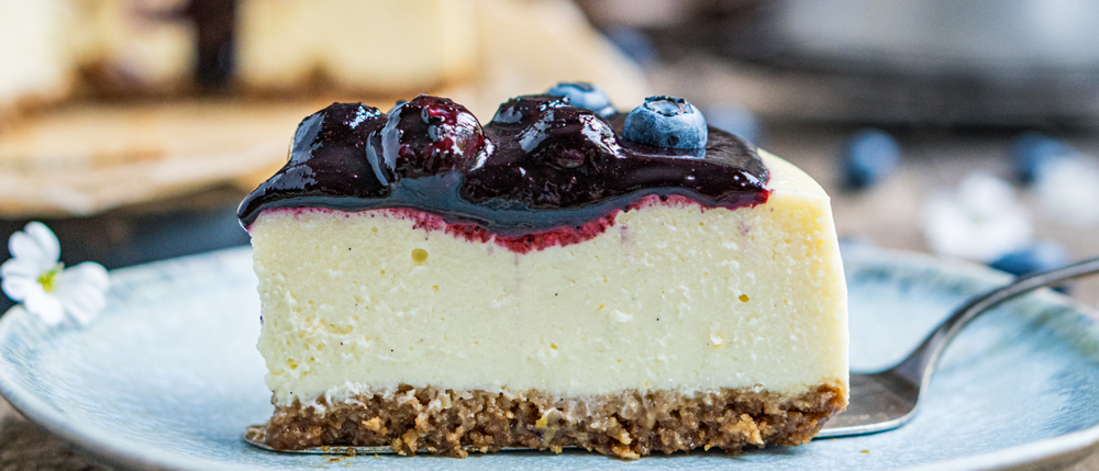 Ein Stück Käsekuchen mit Keksboden und dunkler Beerensoße, garniert mit Blaubeeren, auf einem blauen Teller mit Löffel.