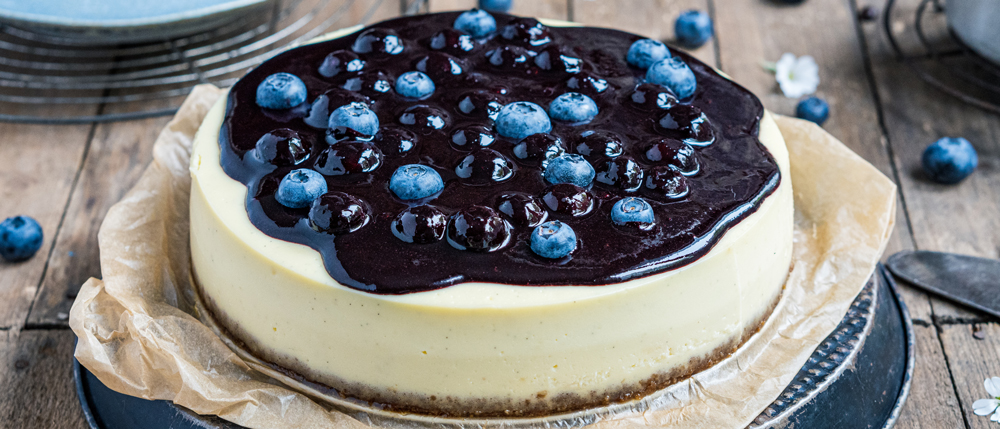 Käsekuchen mit glänzender Blaubeer-Fruchtschicht und frischen Blaubeeren auf Backpapier und einem rustikalen Holzuntergrund.