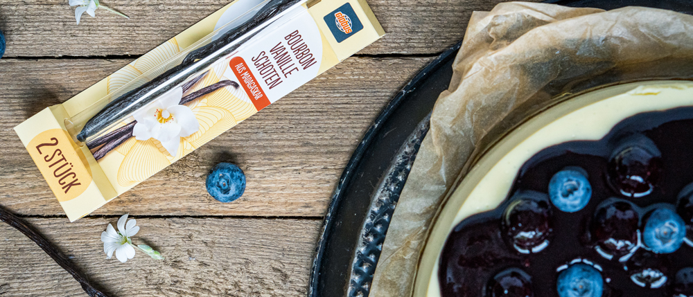 Eine gelbe Packung Bourbon Vanilleschoten neben einem Blaubeerkuchen mit Glasur und frischen Blaubeeren auf Holzuntergrund.