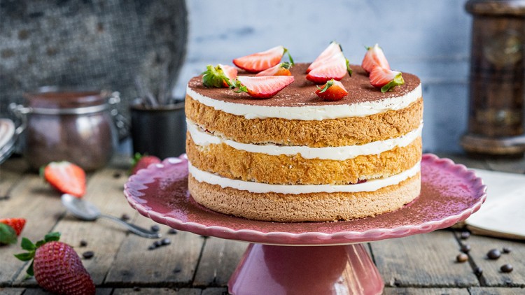 Eine dreischichtige Torte mit heller Creme und Erdbeerstücken als Dekoration steht auf einer rosa Tortenplatte aus Keramik.