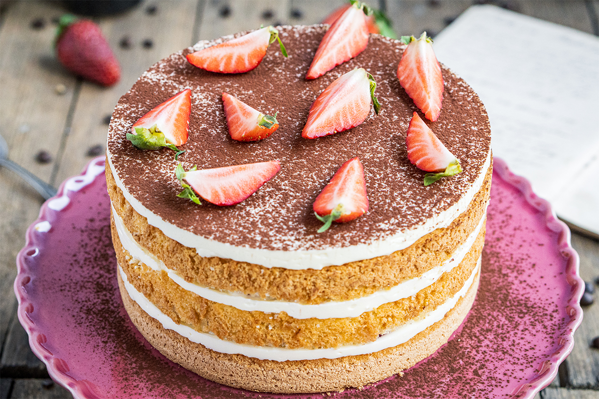 Dreischicht-Torte mit heller Creme, Kakaopulver bestäubt und mit halbierten Erdbeeren dekoriert auf rosa Teller.