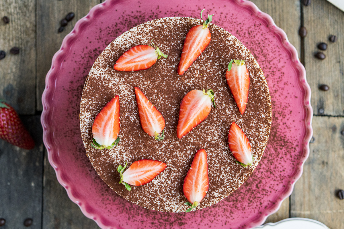 Runde Torte mit Kakao bestäubt, garniert mit halbierten Erdbeeren auf rosafarbenem Teller, Holztisch im Hintergrund.