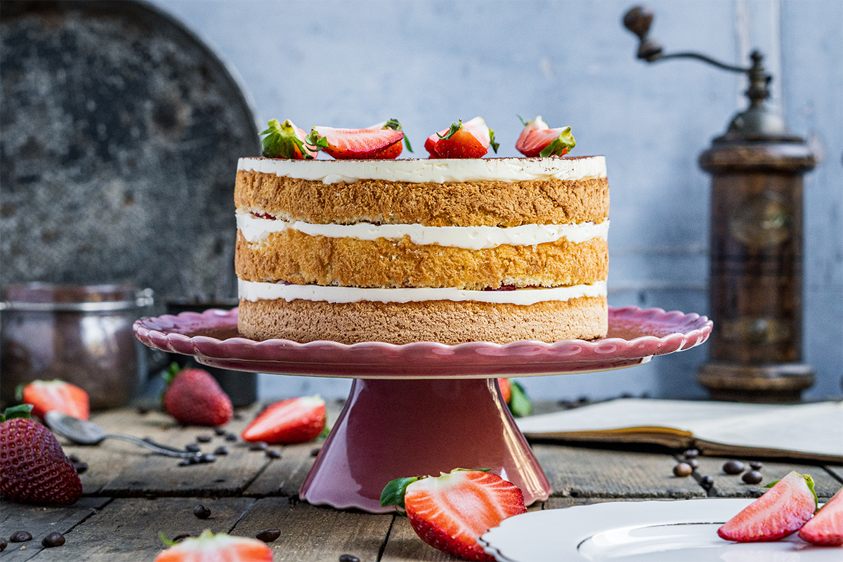 Eine dreischichtige Torte mit Sahne und Erdbeeren auf einer rosa Tortenplatte, dekoriert mit halbierten Erdbeeren.