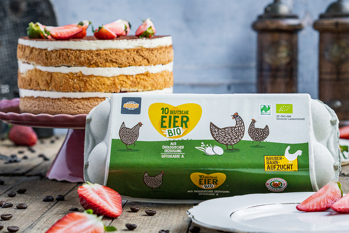 Eine Sahnetorte mit Erdbeeren steht hinter einer Packung GLOBUS Bio-Eier, die mit Hühnern und gelbem Bruderhahn-Aufzucht-Logo bedruckt ist.