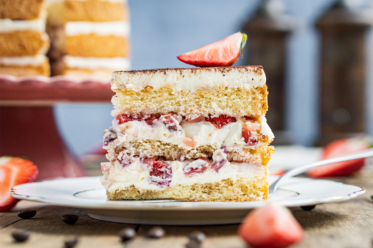 Ein Stück Erdbeer-Sahne-Torte mit hellen Biskuitböden liegt auf einem weißen Teller, dekoriert mit einer halben Erdbeere.