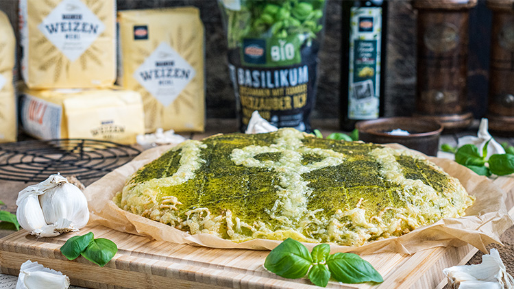 Ein goldbraunes Brot mit grüner Kräuterkruste liegt auf Backpapier, umgeben von frischem Basilikum, Knoblauch und Zutaten.