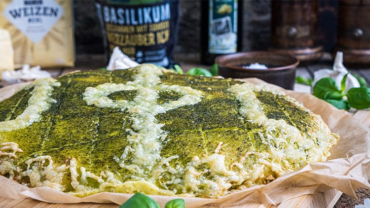 Vegetarische Pizzavariante mit Pesto-Basilikum-Belag auf braunem Backpapier, umgeben von Basilikumblättern und Zutaten.