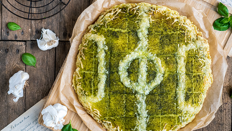Runde, goldbraune Focaccia mit grünem Pesto und einer eingeritzten geometrischen Verzierung, dekoriert mit Knoblauch und Basilikum.