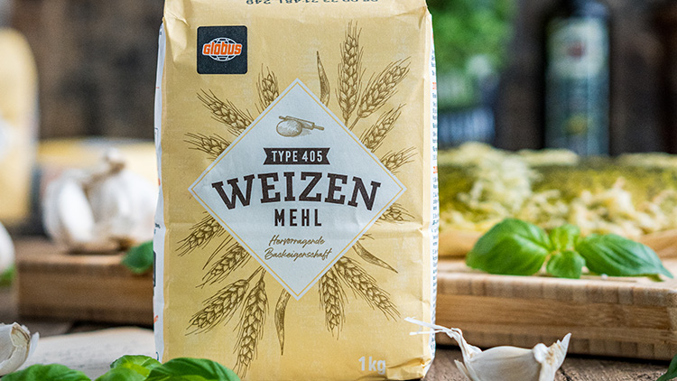 Eine gelbe 1-kg-Packung GLOBUS Weizenmehl Type 405 mit Ährenmuster steht auf einem Holzbrett mit Basilikum und Knoblauch.