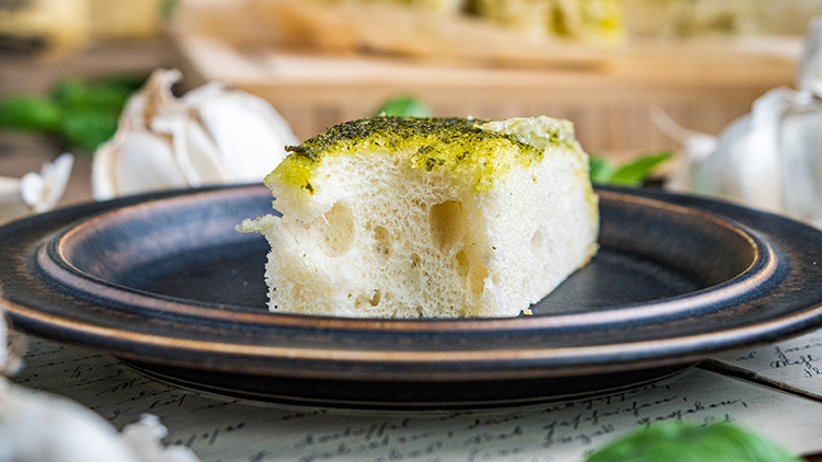 Ein Stück luftig gebackenes Focaccia mit grüner Kräuterkruste auf einem dunklen Teller, im Hintergrund Knoblauch und Basilikum.