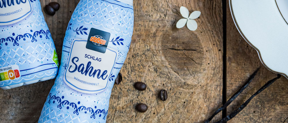 Zwei blaue Sahneflaschen mit GLOBUS-Logo liegen auf Holz, daneben Kaffeebohnen, Vanilleschoten und eine weiße Blüte.