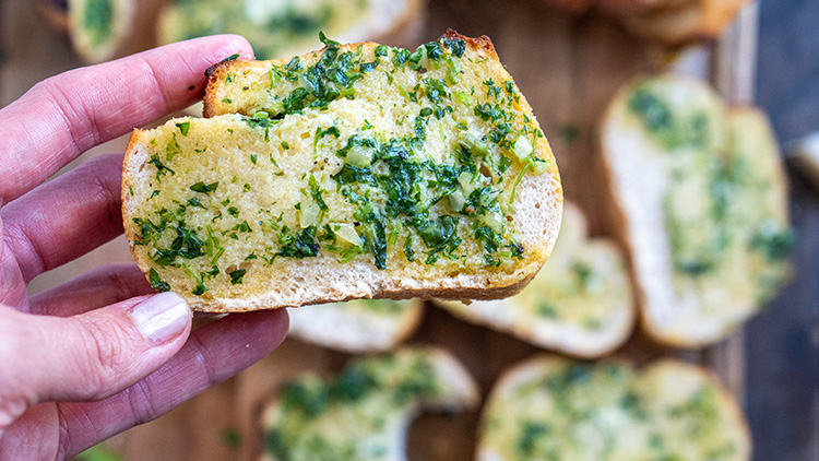 Eine Hand hält ein Stück geröstetes Brot mit gehackten Kräutern und Knoblauch, weitere Brotscheiben im Hintergrund.
