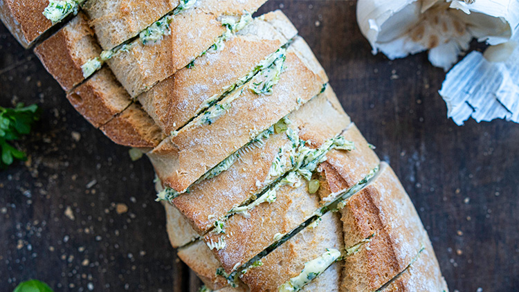 Aufgeschnittenes Brot mit Kräuterbutterfüllung liegt auf einem dunklen Holzuntergrund, daneben Knoblauch und Petersilie. 