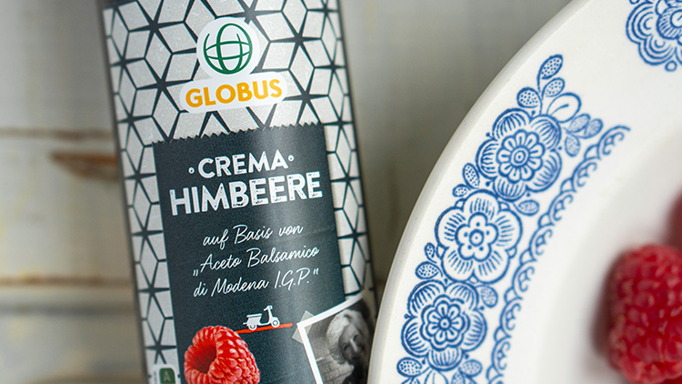 Eine Flasche GLOBUS Crema Himbeere steht neben einem weißen Teller mit blauem Blumenmuster und frischen Himbeeren.