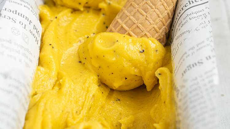 Gelbes Eis mit schwarzen Punkten liegt in einer Manschette aus bedrucktem Papier, daneben eine umgedrehte Eiswaffel.