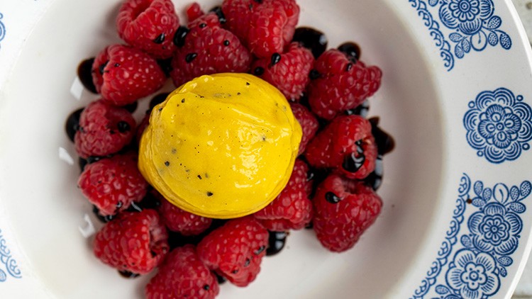 Eine gelbe Eiskugel liegt auf frischen Himbeeren, umgeben von dunklen Balsamico-Tropfen auf einem Teller mit blauem Blumenmuster.