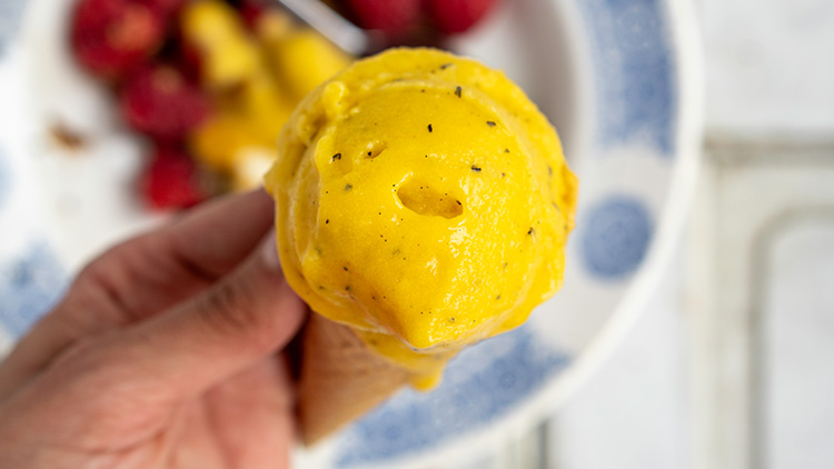Hand hält Eiswaffel mit gelbem Sorbet, im Hintergrund verschwommen Himbeeren und gelbe Stücke auf weiß-blauem Teller.
