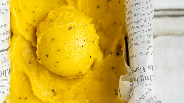 Eine Kugel gelbes Eis mit schwarzen Punkten liegt auf gelbem Eis in einem mit Zeitungspapier ausgelegten Behälter.